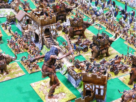 View from behind a wooden palisade with towers and a giant, chariots, war mammoths and Barbarian infantry