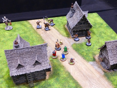 A small wargaming table with three houses and a band of adventurers running into a group of monstrous creatures