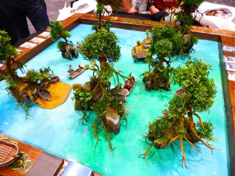 A small gaming table with little islands in a crystal blue lagoon and fantasy figures fighting around row boats