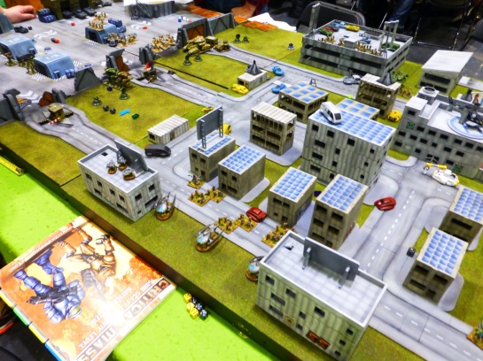 A wargaming table with a model city of high rise buildings and various vehicles and stands of infantry 
