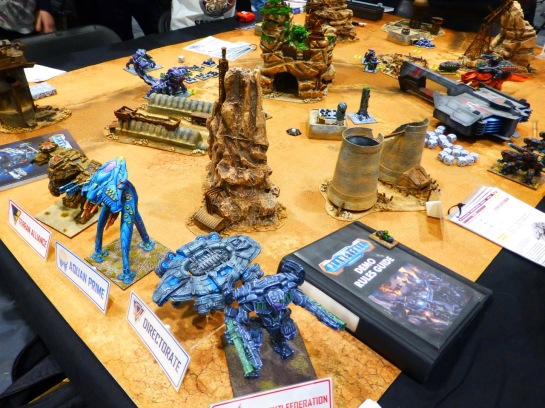 A wargaming table with desert terrain and abandoned industrial buildings and various large mech style war machines