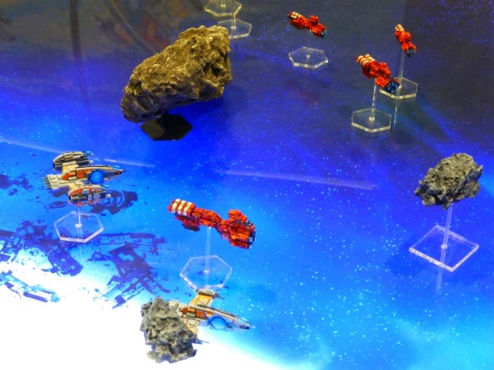 Red and grey space ships amongst asteroids on an underlit blue gaming table