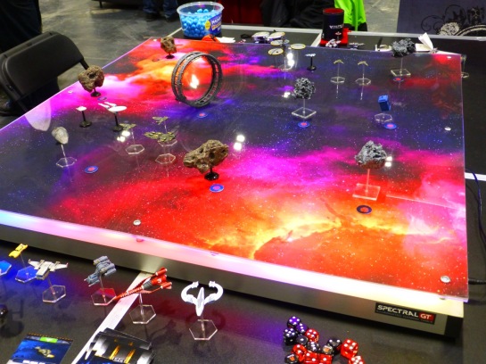 A gaming table showing a backdrop of dark space and red nebulas lit from underneath and with a variety of spacecraft and asteroids placed on top