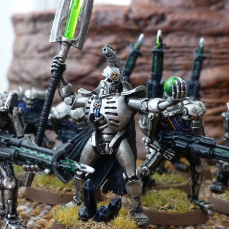 Metal skeletal robot warrior holding a green orb in his left and a staff with a green rod in his right, surrounded by robots with rifles
