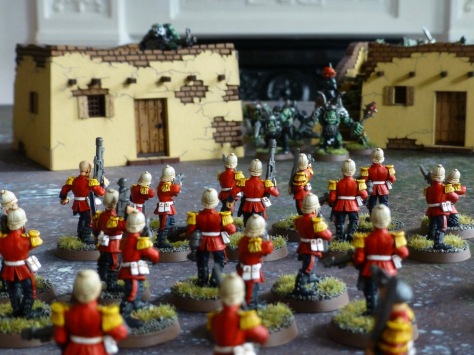 Ranks of soldiers in red jackets and tropical helmets advancing on two adobe buildings with Orks in the background